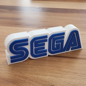 May include: White 3D printed SEGA logo with blue lettering. The letters are stacked together and have a layered effect.
