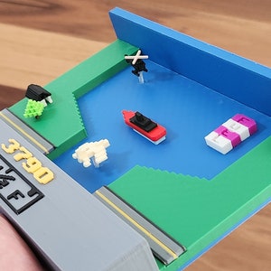 Puede incluir: Un modelo en miniatura impreso en 3D de una escena de videojuego. La escena presenta un barco rojo, un helicóptero, un faro y árboles. El modelo está sobre una base gris con el texto "3790 E 1/4 F" impreso en él.