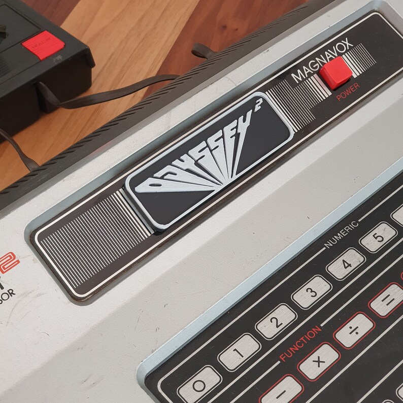May include: A silver and black Magnavox Odyssey 2 video game console with a black and white logo and a red power button. The console has a numeric keypad with black buttons and red labels.