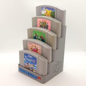 May include: A gray plastic stand for Nintendo 64 game cartridges. The stand holds five cartridges, with the top cartridge showing a picture of Pikachu from the Pokemon video game. The stand has the Nintendo 64 logo on the front.