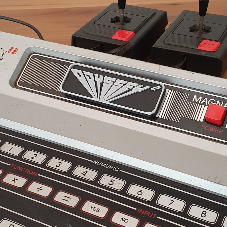 May include: A vintage Odyssey 2 Magnavox video game console with two controllers, a power button, and a numeric keypad. The console is silver and black with a black and silver Odyssey 2 logo.
