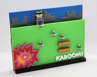 May include: A 3D printed scene depicting a pixelated character in a green and black striped shirt and a green hat standing on a green platform. The character is holding a bomb. The platform is surrounded by a blue sky with gray buildings. There are several bombs exploding on the platform. The text "KABOOM!" is written in white letters on the platform.
