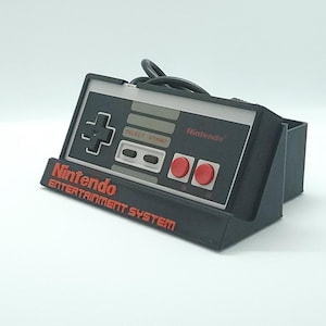 Op de afbeelding: Een zwarte Nintendo Entertainment System-controller met rode knoppen en een grijze frontplaat. De controller staat in een zwarte standaard met het Nintendo-logo aan de voorkant.