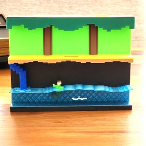 May include: A 3D printed model of a video game level with a green background, brown trees, a blue waterfall, and a blue water surface with a green figure standing on a blue wave. The model is on a black base with the text "Retro Pixel" printed on it.