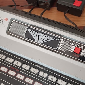 May include: A vintage Magnavox Odyssey 2 video game console with a black and silver design. The console has a red power button and a numeric keypad with function keys. The console has the text "ODYSSEY 2" and "MAGNAVOX" printed on it.