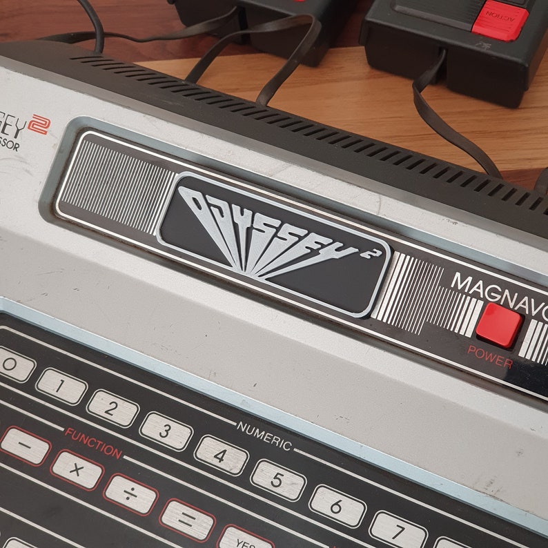 May include: A vintage Magnavox Odyssey 2 video game console with a black and silver design. The console has a red power button and a numeric keypad with function keys. The console has the text "ODYSSEY 2" and "MAGNAVOX" printed on it.