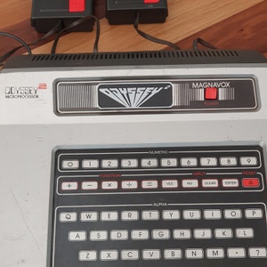 May include: A vintage Magnavox Odyssey 2 video game console with a black and silver design. The console has a built-in keyboard with numeric and alphabetic keys, as well as function keys, input keys, and a reset button. The console also has a power button and a red LED light.