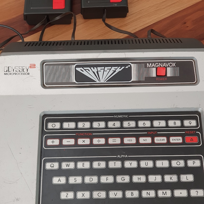 May include: A vintage Magnavox Odyssey 2 video game console with a black and silver design. The console has a built-in keyboard with numeric and alphabetic keys, as well as function keys, input keys, and a reset button. The console also has a power button and a red LED light.
