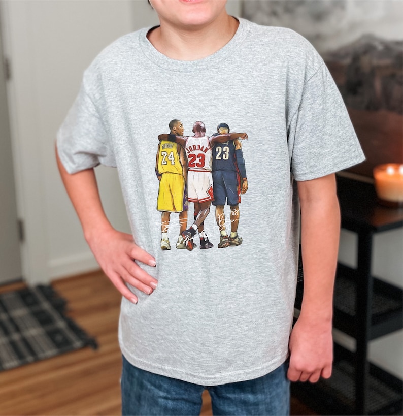 May include: A light gray t-shirt featuring a graphic of three basketball players with the numbers 24, 23, and 23 on their jerseys. The players are in yellow, red, and blue uniforms. The shirt is made of a soft material.