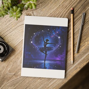 May include: A white notebook featuring a rectangular print of a silhouette of a ballerina dancing against a dark blue cosmic background with swirling purple nebula clouds and bright white stars. The notebook is positioned on a wooden surface.