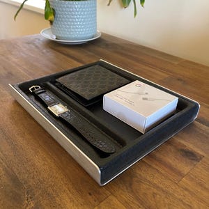 May include: A silver tray with a black felt lining holds a black leather wallet, a wristwatch with a silver face and black leather band, and a white box with the Google logo and the text "USB-C to USB-A Cable 1m (3.3ft)".
