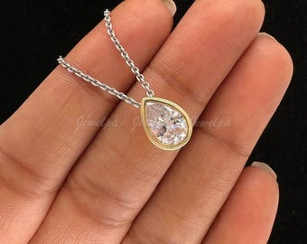 May include: A pear-shaped pendant necklace with a clear gemstone set in a gold bezel. The pendant hangs from a delicate silver chain. The necklace is displayed against a dark background.