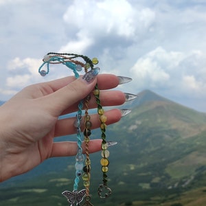 May include: Three beaded bracelets with charms, including a butterfly and a leaf, against a backdrop of a mountain range and a cloudy sky.