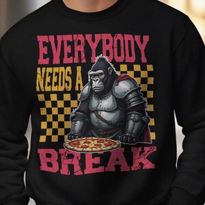 以下が含まれることがあります： ピザを持つ鎧を着たゴリラのグラフィックがプリントされた黒いスウェットシャツ。「EVERYBODY NEEDS A BREAK」の文字が赤と黄色でプリントされています。デザインには市松模様が含まれています。