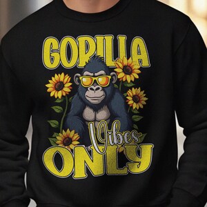 以下が含まれることがあります： サングラスをかけたゴリラのグラフィックが特徴の黒いスウェットシャツ。ひまわりと「GORILLA Vibes ONLY」の黄色い文字がデザインされています。カジュアルウェアに適した漫画風のデザインです。