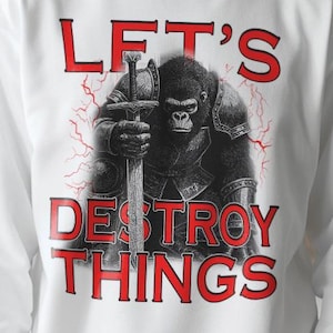 以下が含まれることがあります： 白いスウェットシャツ。剣を持つゴリラ戦士のイラストと、赤い文字で「LET'S DESTROY THINGS」と書かれています。デザインには稲妻のアクセントが含まれており、大胆でエッジの効いた美学を作り出しています。