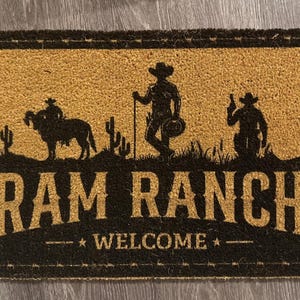 May include: A rectangular doormat with a brown background and a black border. The mat features a silhouette of a cowboy riding a ram, cacti, and the words "RAM RANCH WELCOME".