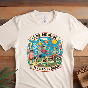 May include: White t-shirt with a colorful graphic of children playing. The graphic includes a child riding a bicycle, a child flying a kite, and a child holding a ball. The text "Leave Me Alone My Dad Is Dead" is written in a banner around the graphic.