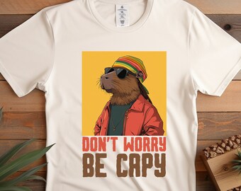 May include: Off-white t-shirt featuring a capybara wearing sunglasses, a colorful hat, and a red jacket. The shirt has the text "DON'T WORRY BE CAPY" in brown and red letters. The design is on a yellow background.