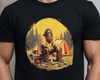Può includere: Una maglietta nera con una grafica di un Bigfoot seduto vicino a un falò con una tenda sullo sfondo.