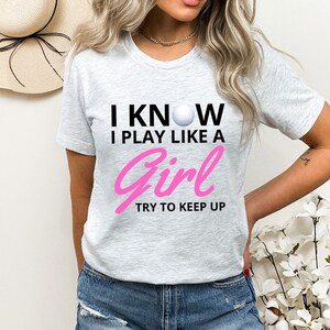 May include: Gray t-shirt with the text "I know I play like a girl try to keep up" in black and pink. A white golf ball is in the text.