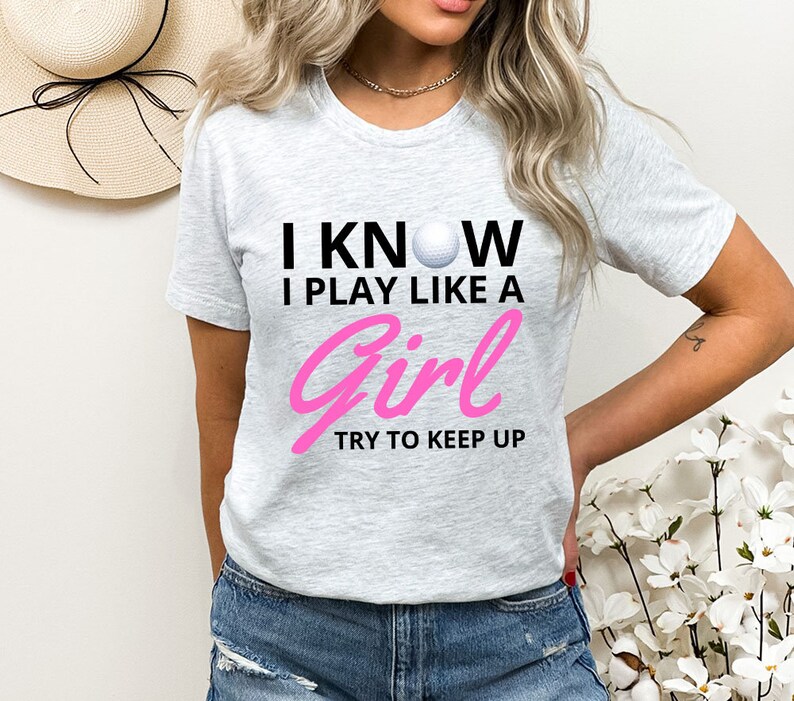 May include: Gray t-shirt with the text "I know I play like a girl try to keep up" in black and pink. A white golf ball is in the text.