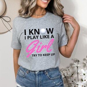 May include: Gray short-sleeved t-shirt with black text that reads "I KNOW I PLAY LIKE A GIRL TRY TO KEEP UP" and a white golf ball graphic. The text "GIRL" is in pink.