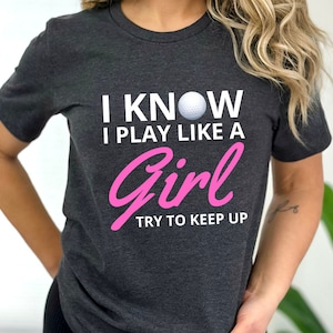May include: A dark gray t-shirt with the text "I know I play like a girl try to keep up" in white and pink lettering. A white golf ball is centered above the text.
