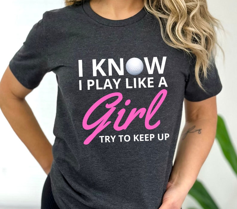 May include: A dark gray t-shirt with the text "I know I play like a girl try to keep up" in white and pink lettering. A white golf ball is centered above the text.