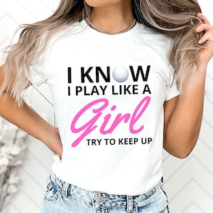May include: White t-shirt with black text that reads "I know I play like a girl try to keep up". The text is in a bold font. There is a pink golf ball graphic above the text.