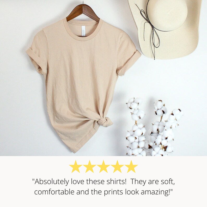 May include: A beige t-shirt with rolled up sleeves is hanging on a wooden hanger. The shirt is knotted at the bottom. The t-shirt is on a white background with a straw hat and a cotton plant.