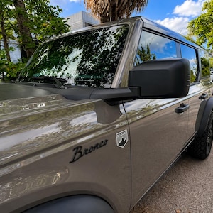 May include: A gray SUV with black accents, parked outdoors. The word "Bronco" is displayed in black script on the side. A black emblem with a mountain and tree design is visible. The vehicle has black tires and a black side mirror.