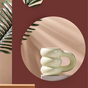 Könnte beinhalten: Eine hellgrüne Keramiktasse mit einem einzigartigen, abgerundeten, strukturierten Design und einem großen Henkel. Die Tasse steht auf einer weißen Plattform vor einem braunen Hintergrund.