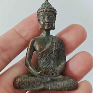 Puede incluir: Pequeña estatua de Buda de bronce en posición de meditación sentado. La estatua tiene una túnica detallada y una expresión serena. El bronce tiene una pátina, dándole un aspecto envejecido. La estatua se sostiene en una mano.