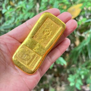 May include: A gold rectangular bar featuring embossed Chinese characters and designs. The bar is held in a hand, set against a blurred green backdrop. The gold bar has a shiny, reflective surface.