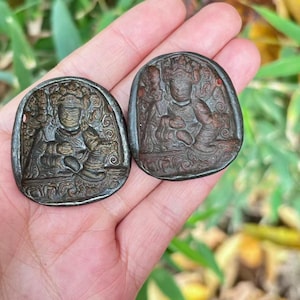 May include: Two antique, oval-shaped bronze pendants. Each pendant features a detailed relief carving of a seated figure wearing a crown and ornate clothing. The figures are surrounded by intricate patterns and designs. The pendants have a dark, aged patina.