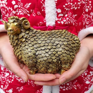 Puede incluir: Escultura de carnero de color bronce con detalles en forma de monedas. El carnero tiene cuernos curvados y se muestra sobre un fondo rojo y blanco estampado. Objeto decorativo.