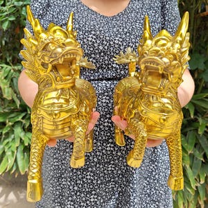 Może przedstawiać: Dwa złote, bogato zdobione posągi mitycznych stworzeń, być może smoków lub lwów, są prezentowane. Posągi mają szczegółowe cechy, w tym rogi, otwarte usta i skomplikowane wzory ciała. Posągi są wykonane z błyszczącego metalu.