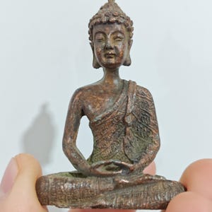 Puede incluir: Pequeña estatua de Buda de color bronce, en posición de meditación sentado. La figura tiene rasgos faciales detallados, una expresión serena y está cubierta con una túnica. La estatua se sostiene en una mano sobre un fondo blanco.