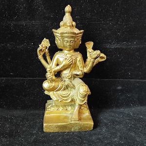 May include: A gold-colored statue of a seated deity with multiple arms and faces. The figure is adorned with ornate headwear and holds various objects in its hands. The statue is on a square base and appears to be made of metal.