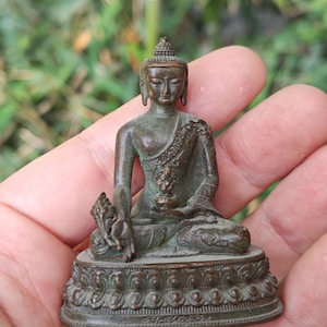 Puede incluir: Pequeña estatua de Buda de color bronce, en posición de meditación sentado. La estatua presenta detalles intrincados, incluyendo una base detallada y ropa ornamentada. La figura se sostiene en una mano sobre un fondo verde borroso.