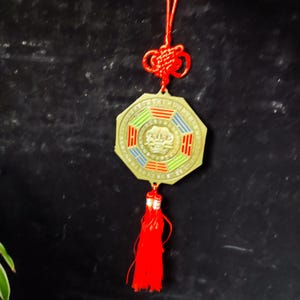 May include: A gold-colored, octagonal pendant with red, green, and blue accents. The pendant has a red tassel and a decorative knot at the top, suspended by a red cord. The design is likely of Asian origin.