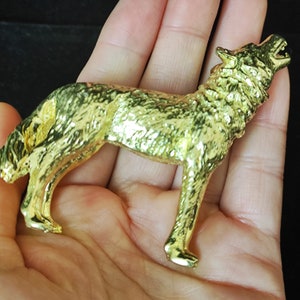 May include: A gold-colored wolf figurine in a howling pose. The wolf is detailed with textured fur and stands on four legs. The figurine is held in a hand against a dark background.