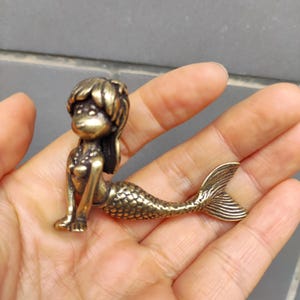 Puede incluir: Pequeña figura de sirena de color bronce. La sirena tiene un diseño estilizado con una cola detallada y cabello suelto. La figura se sostiene en una mano, mostrando su tamaño y detalles intrincados. La estética general es vintage.