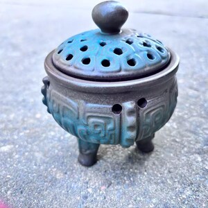 May include: A ceramic incense burner with a teal and gray glaze. The lidded vessel features a perforated top, decorative carvings, and three small feet. The lid has a small, round handle. The incense burner is a decorative item.