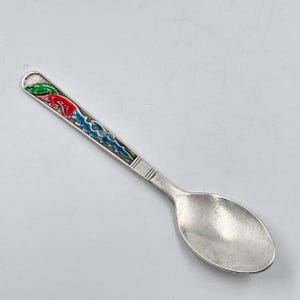 Op de afbeelding: Een zilveren lepel met een kleurrijk emaille ontwerp. De steel heeft een groen, rood en blauw patroon. De lepel heeft een afgeronde kom en een gedetailleerd ontwerp op de steel.