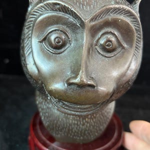 May include: A bronze-colored monkey head sculpture with detailed facial features, including large eyes and a defined nose. The sculpture sits atop a red wooden base, showcasing the monkey's head and upper torso.