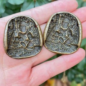 May include: Two antique bronze-colored oval pendants. Each pendant features a detailed relief carving of a seated figure with ornate headdress and clothing. The pendants have a weathered appearance, suggesting age and use.