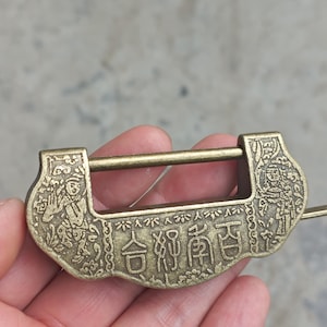 Puede incluir: Un candado de latón antiguo con intrincadas tallas y caracteres chinos. El candado rectangular presenta una barra deslizante y figuras decorativas. El latón tiene un aspecto envejecido y antiguo.