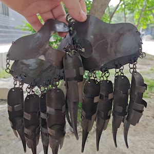 Pu&ograve; includere: Un elemento decorativo in metallo marrone scuro con una serie di elementi appesi e appuntiti. Il pezzo &egrave; tenuto in mano e sembra essere fatto di metallo martellato, con una catena che collega i vari componenti. Il design ha un'estetica unica, forse tribale.